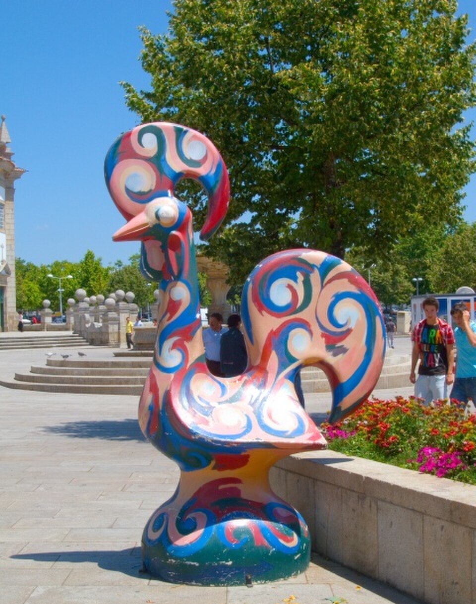 Escultura colorida do Galo de Barcelos em uma praça, com árvores e flores ao fundo.