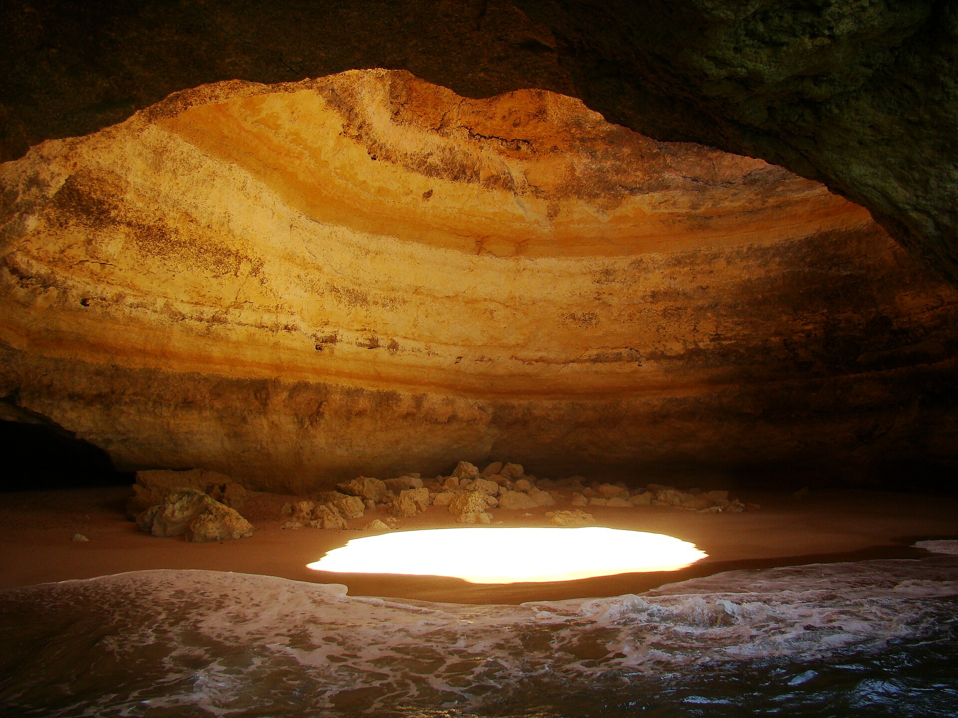 Interior da gruta de Benagil, com abertura no teto rochoso e luz sobre a areia.