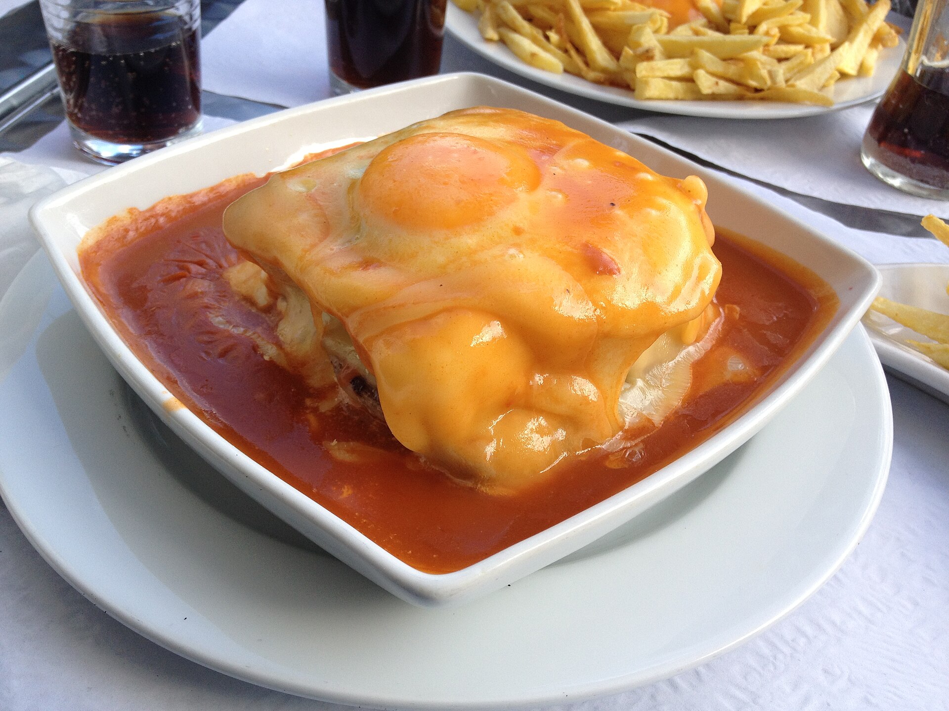 Francesinha coberta com queijo, molho e ovo, acompanhada de batatas fritas.