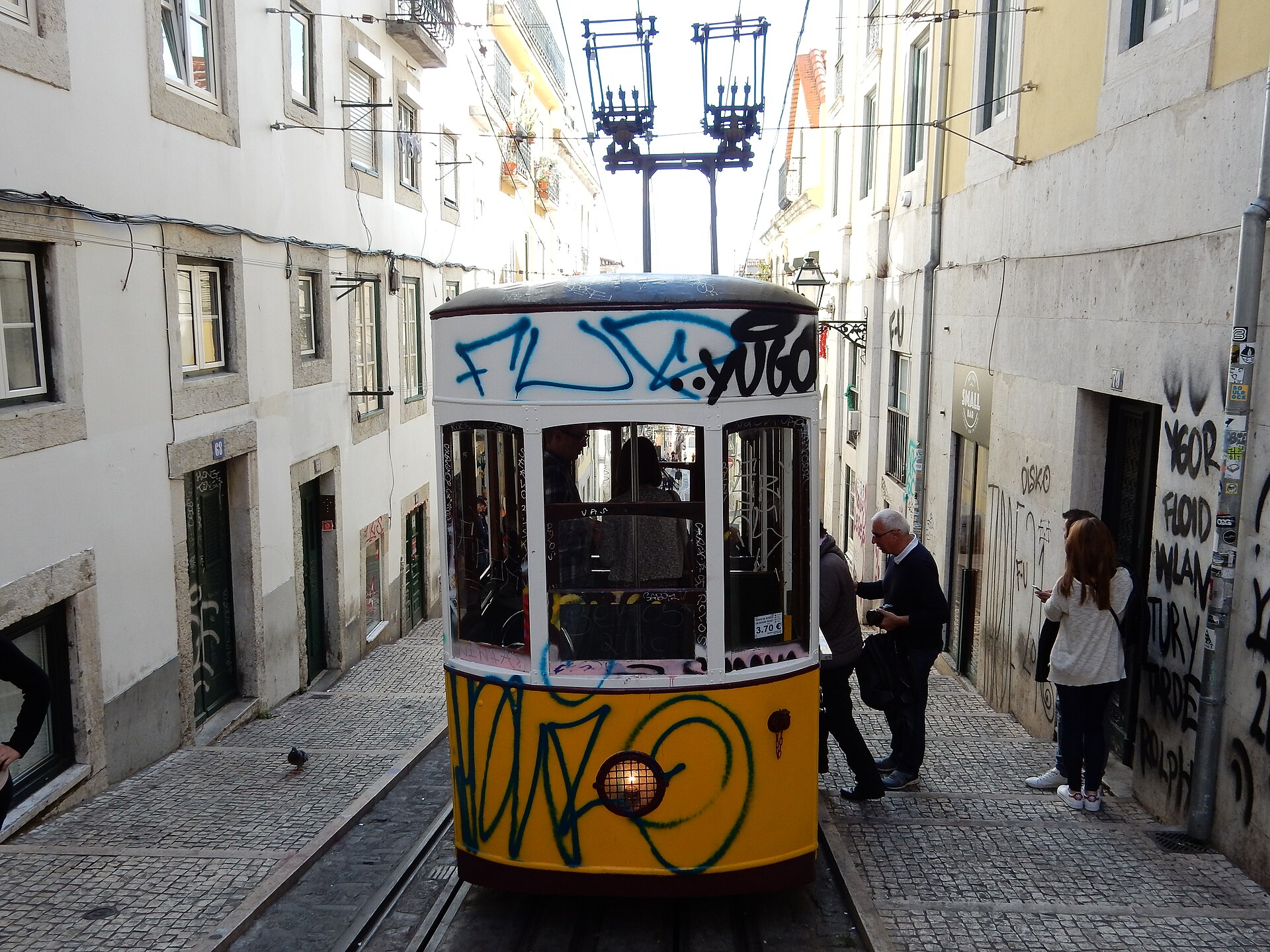 Carruagem amarela do Elevador da Bica entre fachadas e trilhos de uma rua de Lisboa.