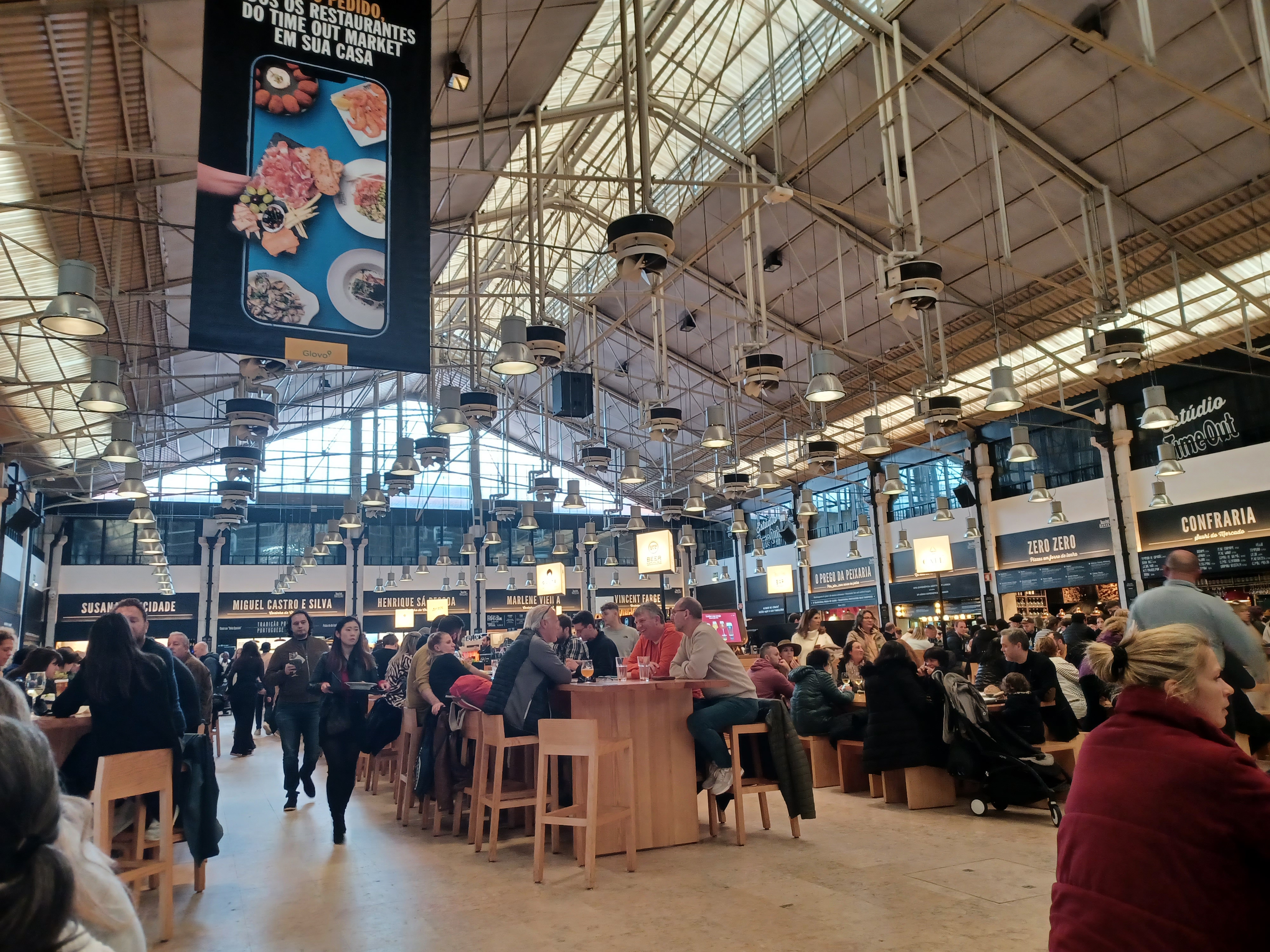 Interior do Mercado da Ribeira, em Lisboa, com bancas de restauração, mesas e visitantes sob a estrutura metálica do salão.