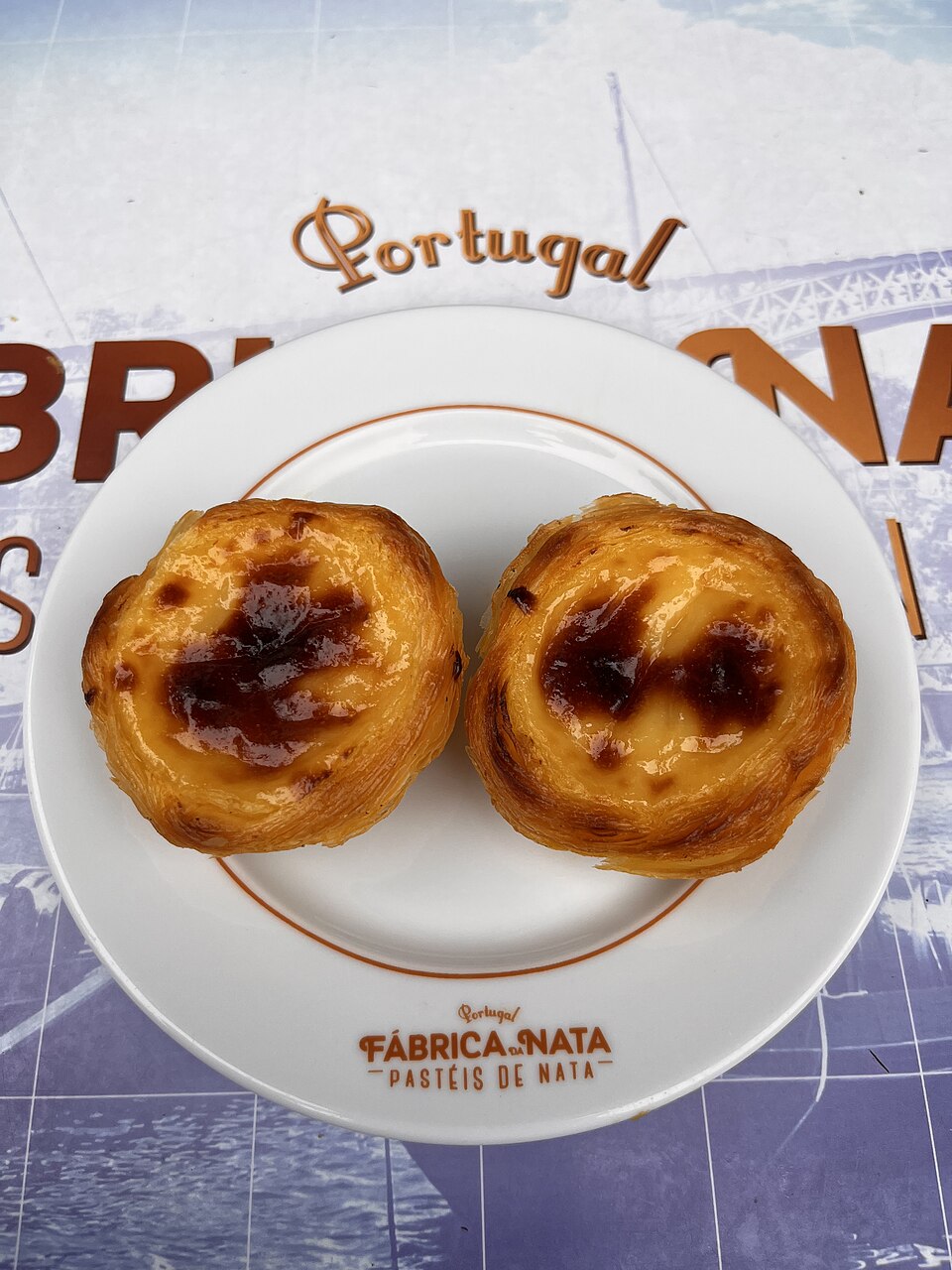 Dois pastéis de nata dourados, servidos em um prato da Fábrica da Nata.